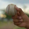 Detailed close-up of a hand holding a baseball outdoors with a blurred background, showcasing grip and texture.